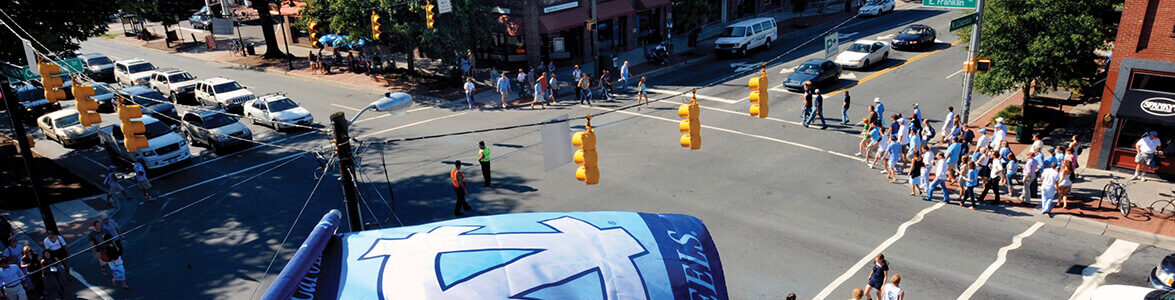 Aerial view of a downtown street crossing on a sunny day with a UNC Tar Heels flag flluttering close up