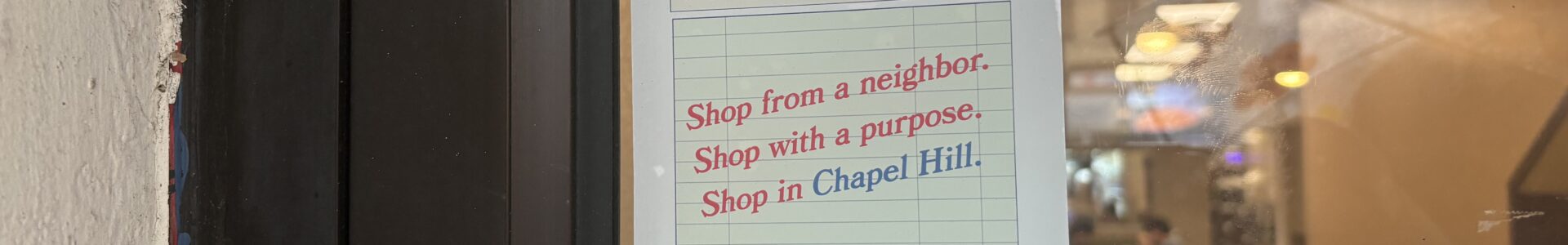 A sticker in the window of a business that reads "Please come again! Shop from a neighbor. Shop with a purpose. Shop in Chapel Hill."